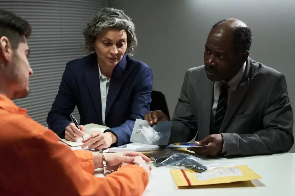 In an interrogation room, two lawyers confer with a handcuffed client. One attorney in Lakeland holds a bag of evidence while the other meticulously takes notes, strategizing their next legal move.
