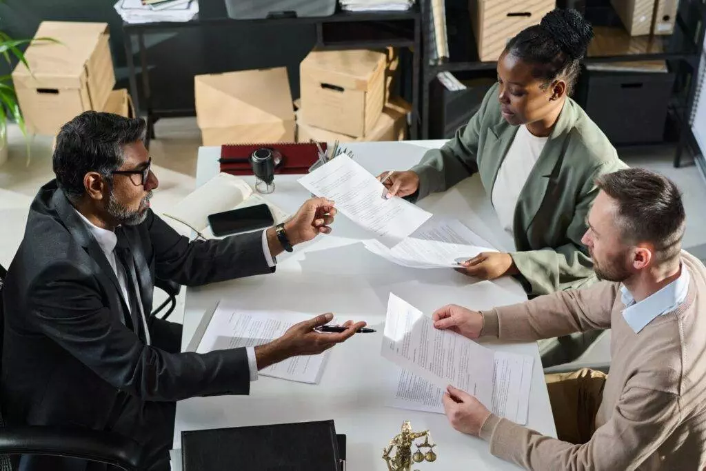 Three people sit around a table in a bustling Florida office, engaged in lively discussion over documents. Surrounded by boxes and office supplies, the setting buzzes with the energy of productivity and warm sunshine seeping through the windows.